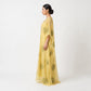 A woman in profile wearing a flowing yellow caftan with green circular motifs, photographed against a plain light gray studio background.