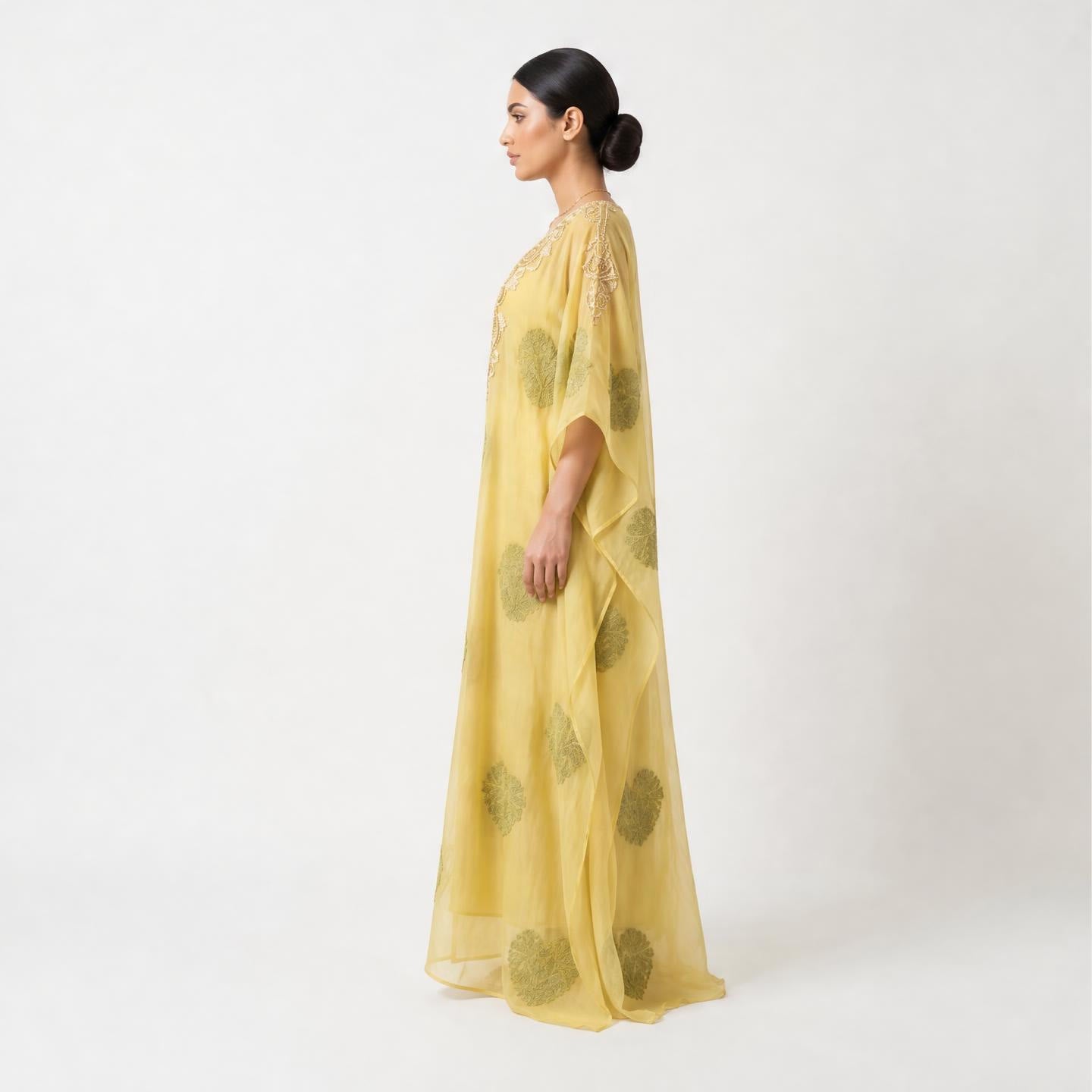 A woman in profile wearing a flowing yellow caftan with green circular motifs, photographed against a plain light gray studio background.