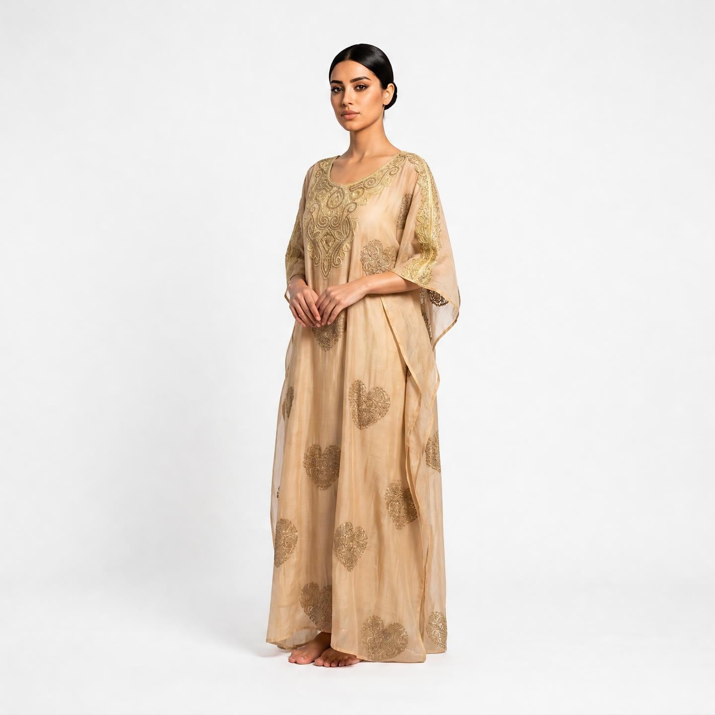 A woman modeling a beige maxi kurta with gold floral embroidery and sheer half sleeves, standing against a plain white studio background.