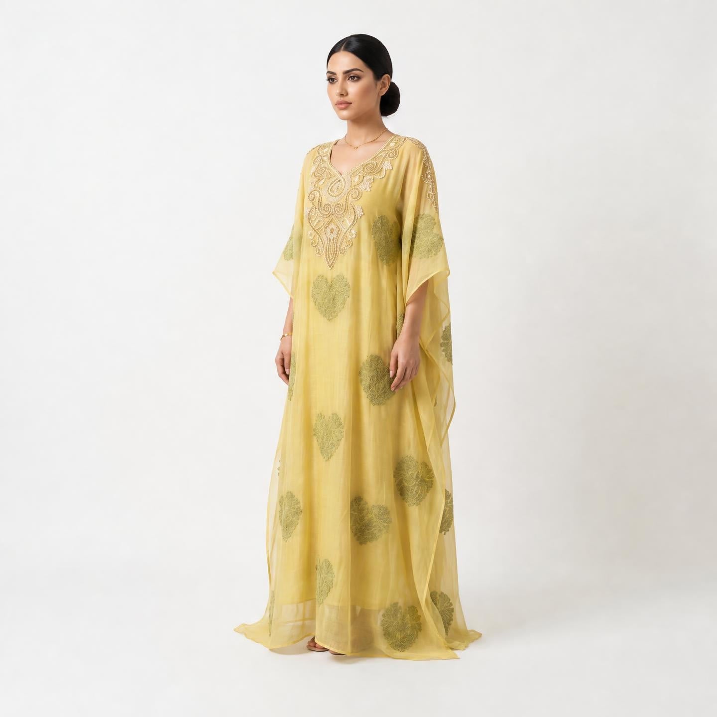 A woman modeling a long, flowing yellow caftan dress with olive green heart patterns and gold embroidery on the neckline, set against a plain white studio background.