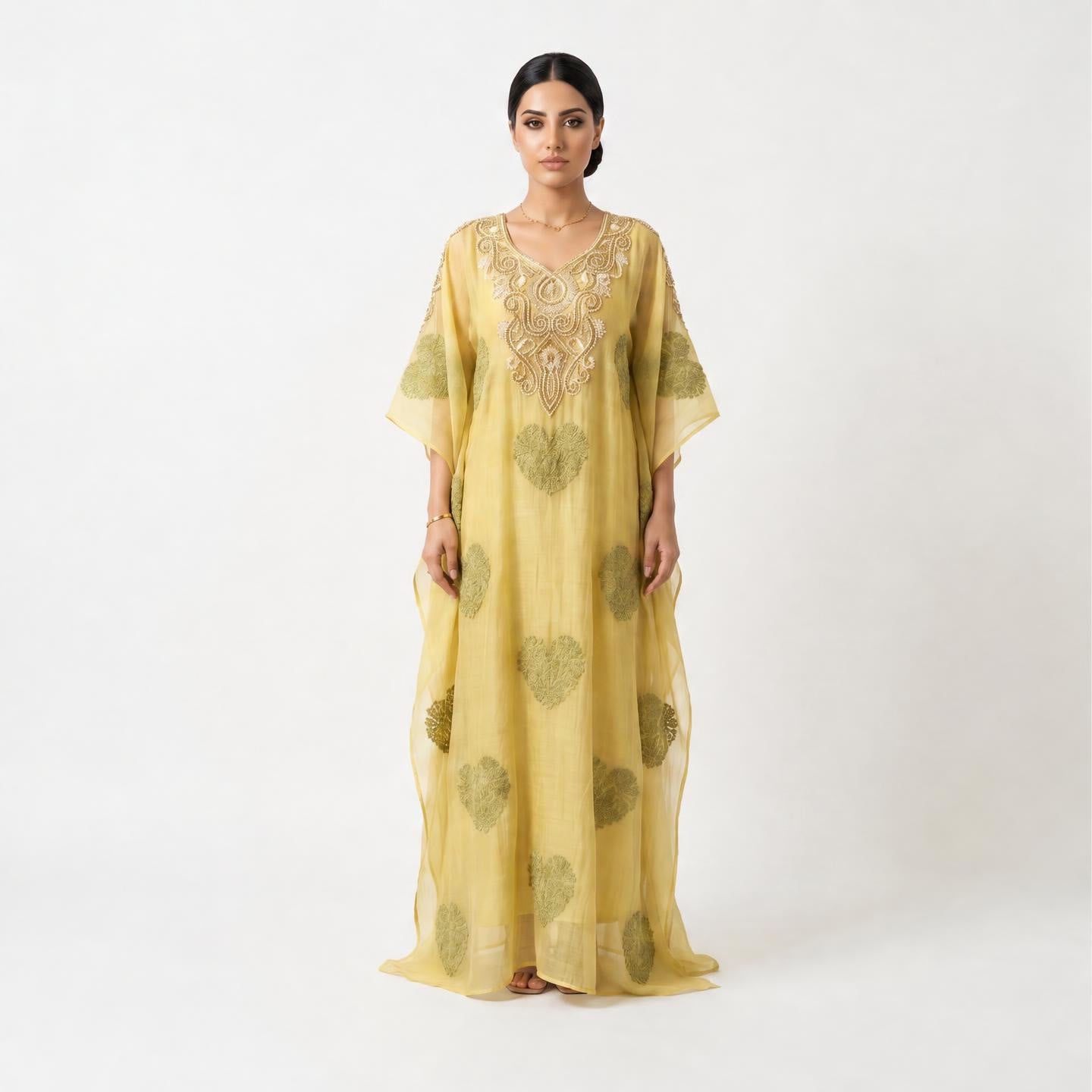 A woman modeling a long, flowing kaftan dress in pale yellow with olive green heart-like motifs and ornate gold embroidery around the neckline, photographed against a plain light gray studio background.