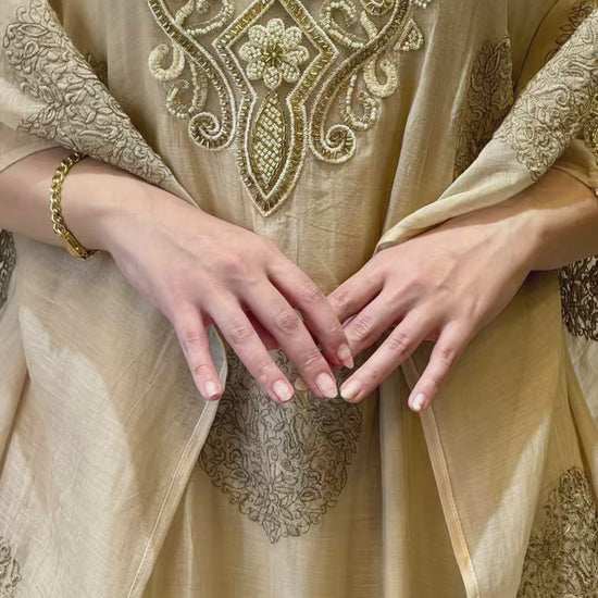 A short aesthetic video loop of the luxury beige 2-piece mul cotton kaftan, showcasing the heavy gold embroidery on the neck and sleeves and the elegant drape of the Armani silk inner in motion.