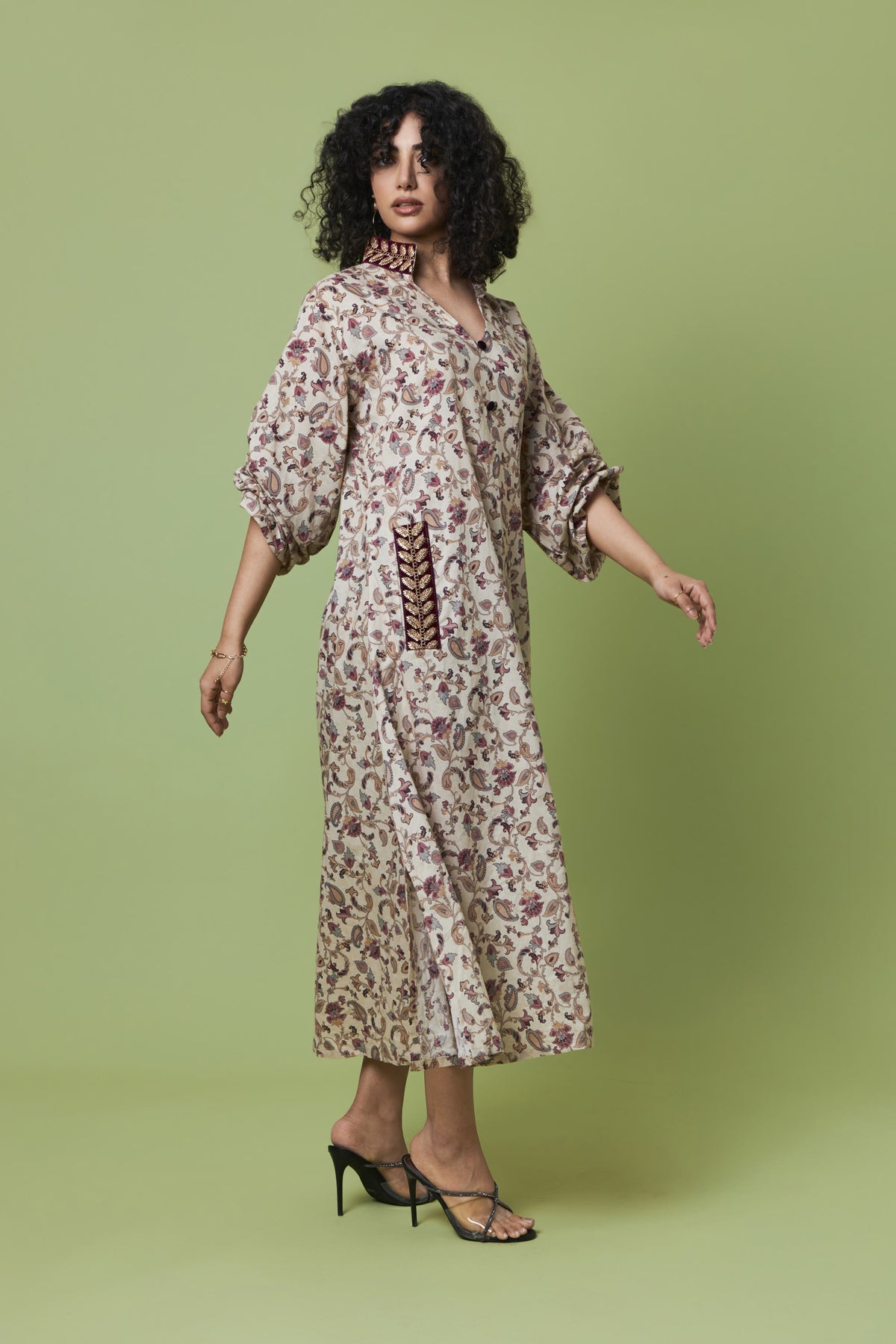 A woman modeling a long-sleeved midi dress with a beige and brown floral pattern, lace-up front detail, and black strappy heels, set against a solid light green studio background.