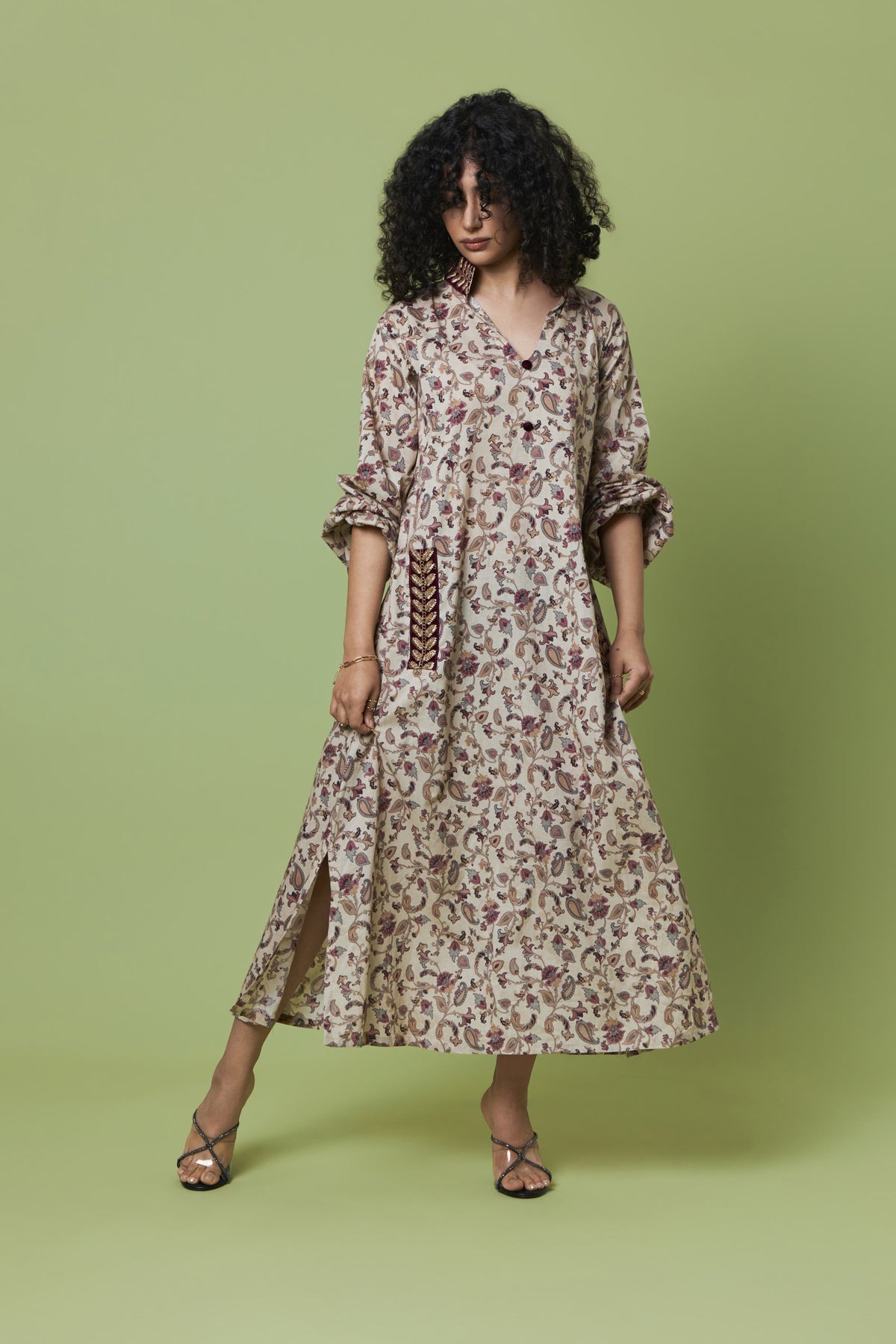 A woman modeling a long-sleeved midi dress with a beige and brown floral pattern, standing against a solid sage green studio background.