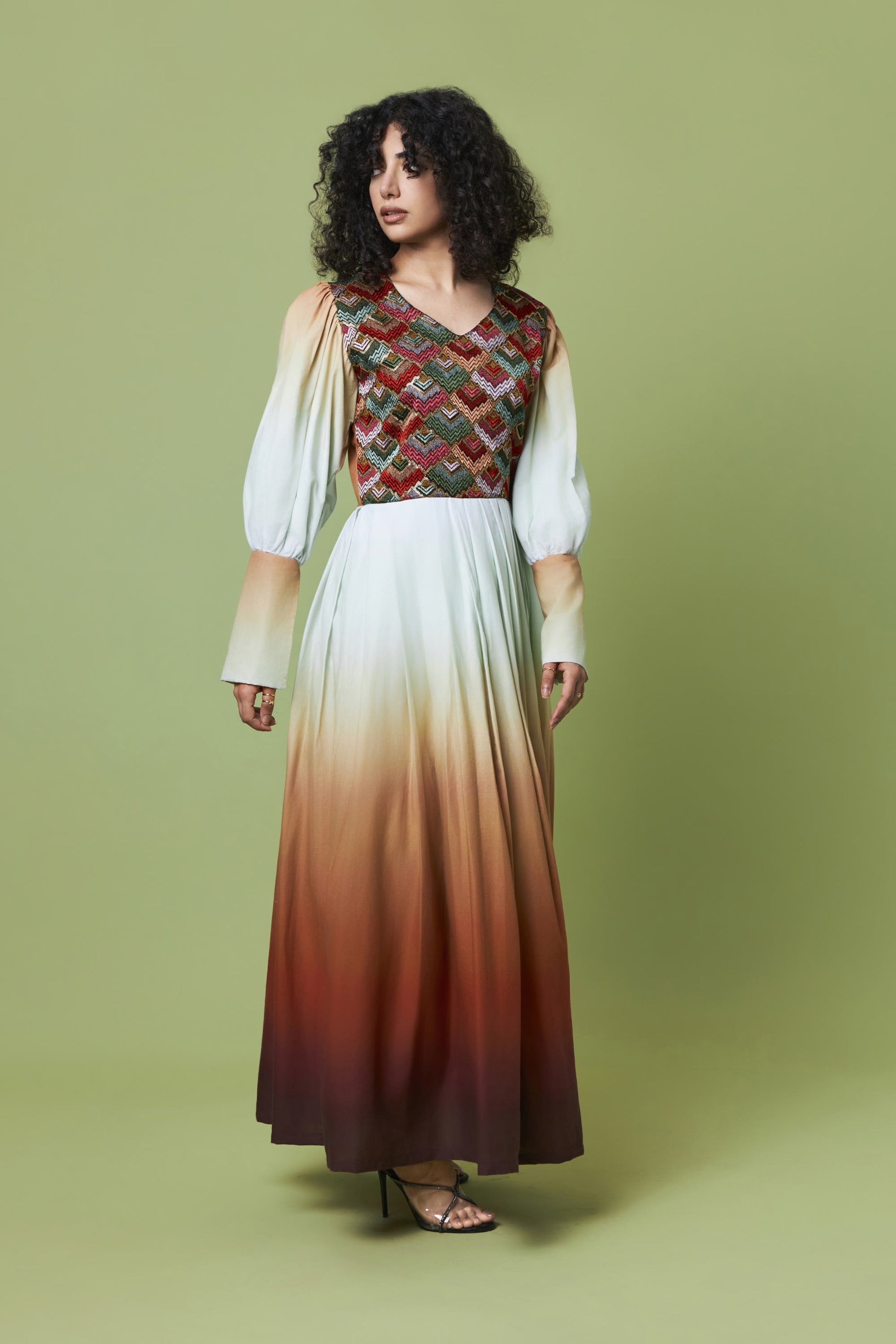 A woman modeling a long maxi dress with a geometric patterned bodice and an ombré skirt fading from cream to rust and dark brown, set against a solid olive green studio background.
