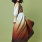 A model with curly black hair is shown from the back wearing a long-sleeved maxi dress with a white-to-brown ombre skirt and a patterned bodice, set against a solid olive green studio background.