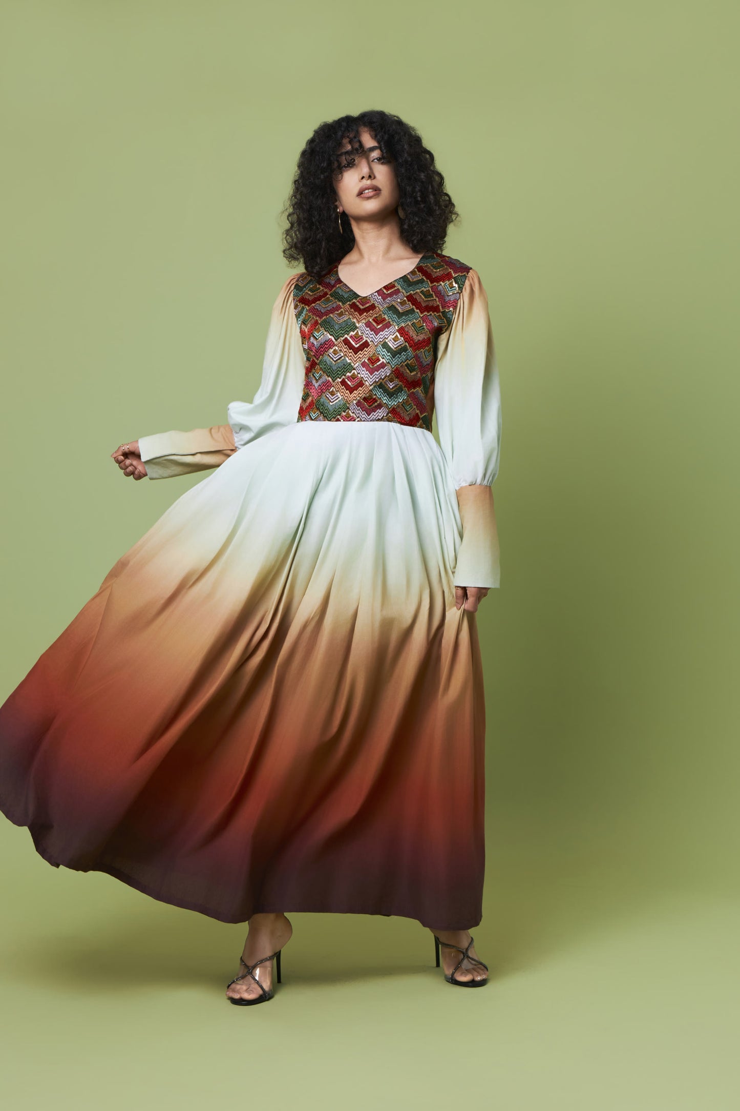 A woman modeling a long-sleeved maxi dress with a multicolor geometric patterned bodice and a flowing white-to-brown ombre skirt, set against a solid sage green studio background.