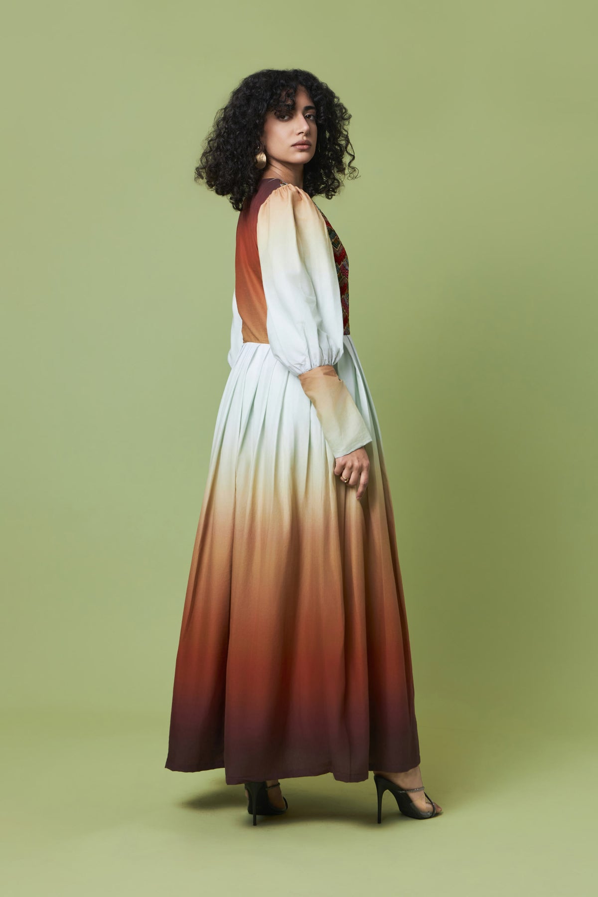 A woman modeling a long-sleeved maxi dress with a vertical gradient design, transitioning from white at the top to rust orange and deep brown at the bottom, set against a solid sage green studio background.
