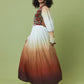 A woman modeling a long maxi dress with a gradient design, featuring a multicolor patterned bodice, white upper skirt, and rust-orange to dark brown ombre lower skirt, set against a solid sage green background.