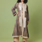 A woman modeling a long, loose-fitting striped tunic or kaftan dress with cream crochet panel details, paired with beige wide-leg pants and black strappy heels, against a solid sage green studio background.
