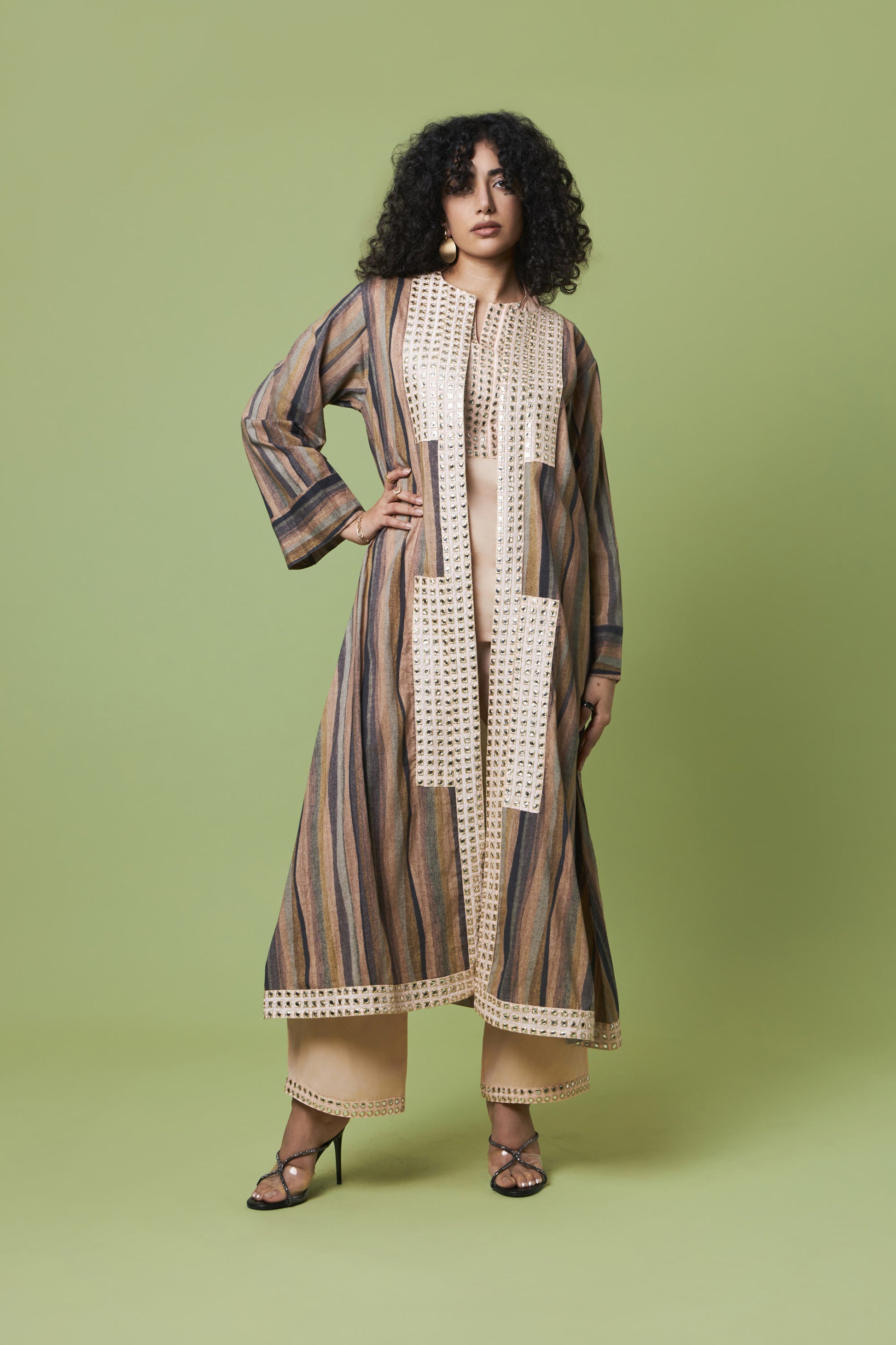 A woman modeling a long, loose-fitting striped tunic or kaftan dress with cream crochet panel details, paired with beige wide-leg pants and black strappy heels, against a solid sage green studio background.