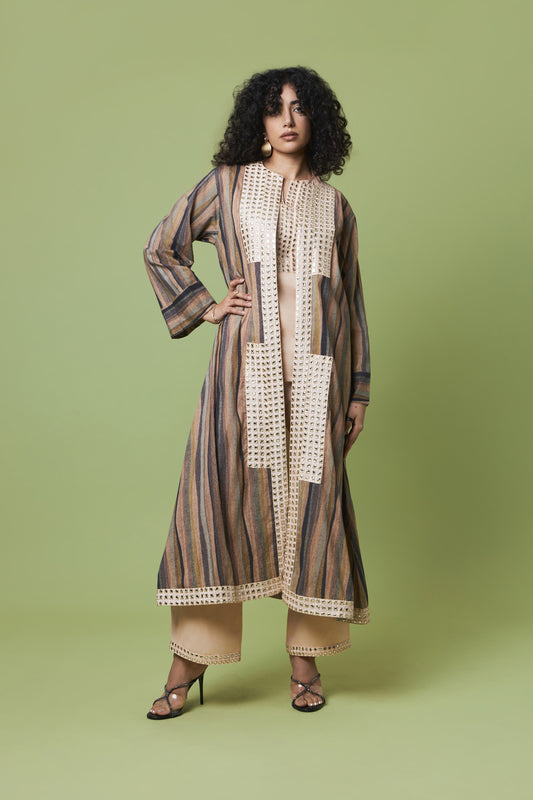 A woman modeling a long, loose-fitting striped tunic or kaftan dress with cream crochet panel details, paired with beige wide-leg pants and black strappy heels, against a solid sage green studio background.