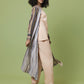 A model with curly black hair is shown from a side-back angle wearing a beige two-piece suit with a striped open-front cardigan and black high heels, set against a solid sage green studio background.