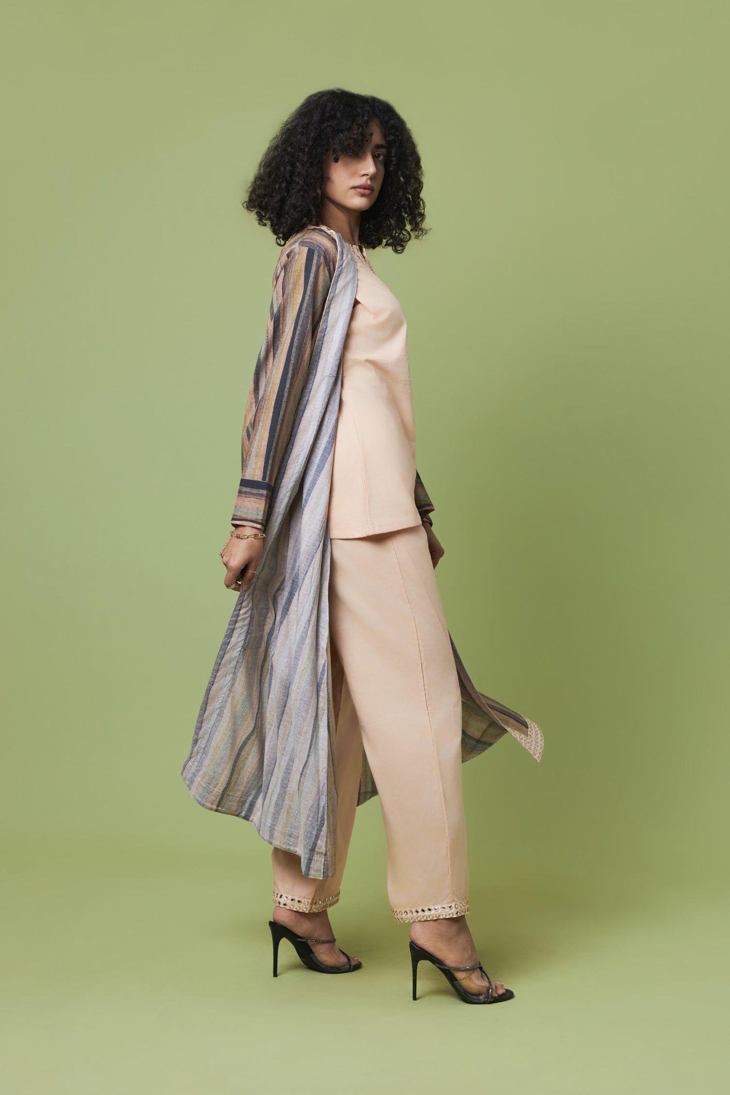 A model with curly black hair is shown from a side-back angle wearing a beige two-piece suit with a striped open-front cardigan and black high heels, set against a solid sage green studio background.