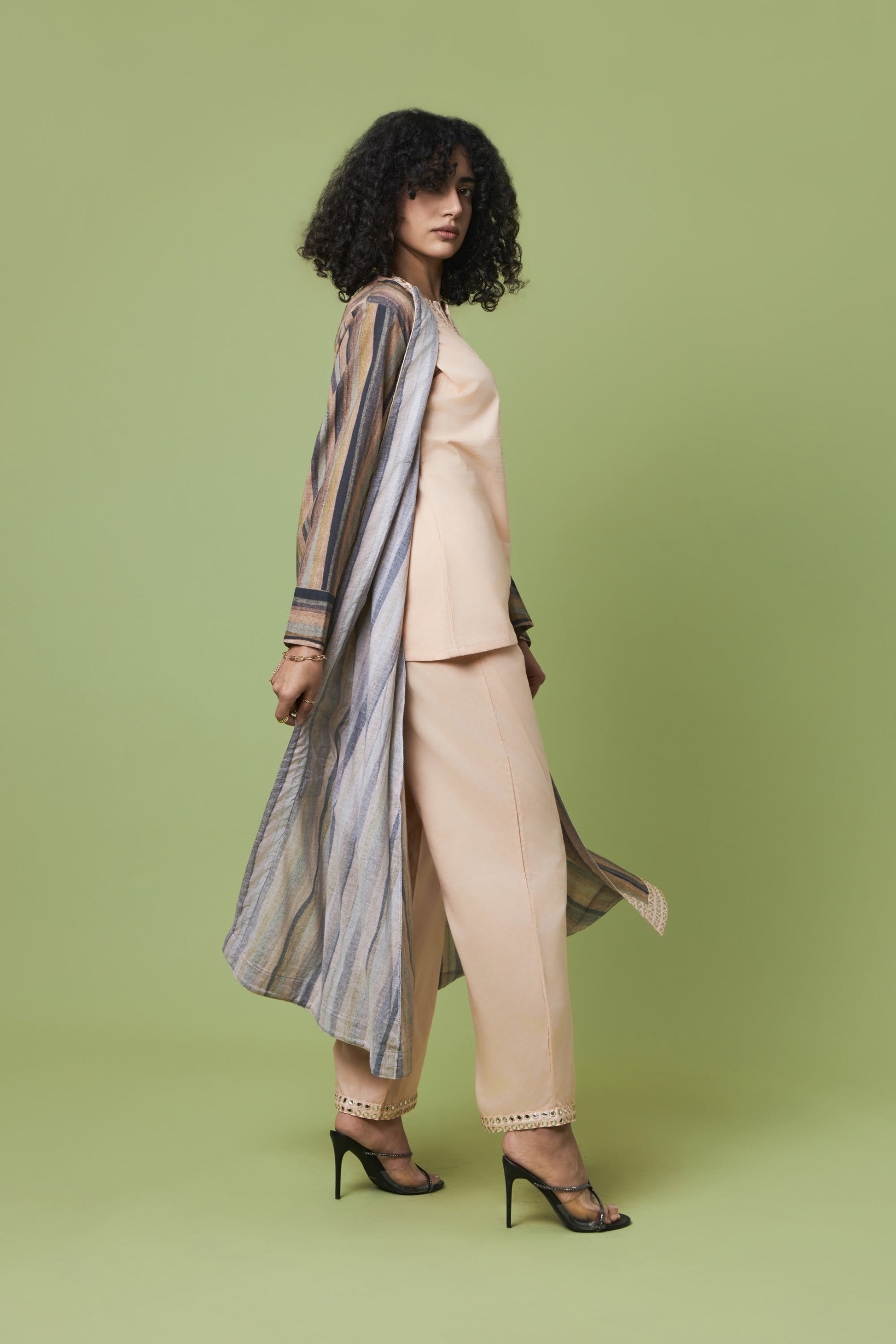 A model with curly black hair is shown from a side-back angle wearing a beige two-piece suit with a striped open-front cardigan and black high heels, set against a solid sage green studio background.