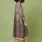 A woman modeling a long, loose striped tunic dress in beige, brown, and muted blue tones, paired with beige pants and black high heels, set against a solid sage green studio background.