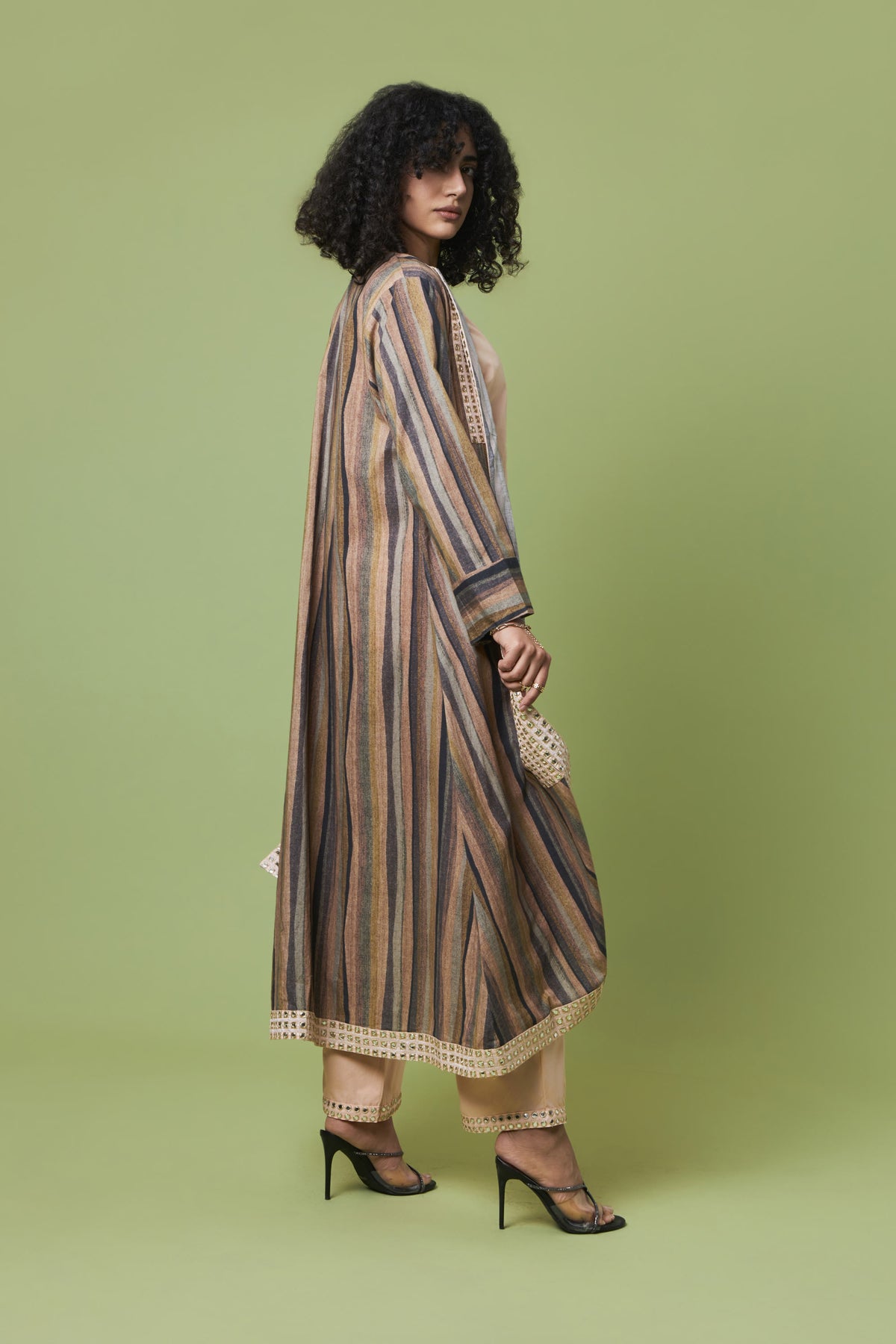 A woman modeling a long, loose striped tunic dress in beige, brown, and muted blue tones, paired with beige pants and black high heels, set against a solid sage green studio background.