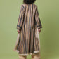 A woman viewed from behind wearing a long beige and brown vertically striped tunic with cream lace trim, paired with beige pants and black heels, against a solid light green studio background.