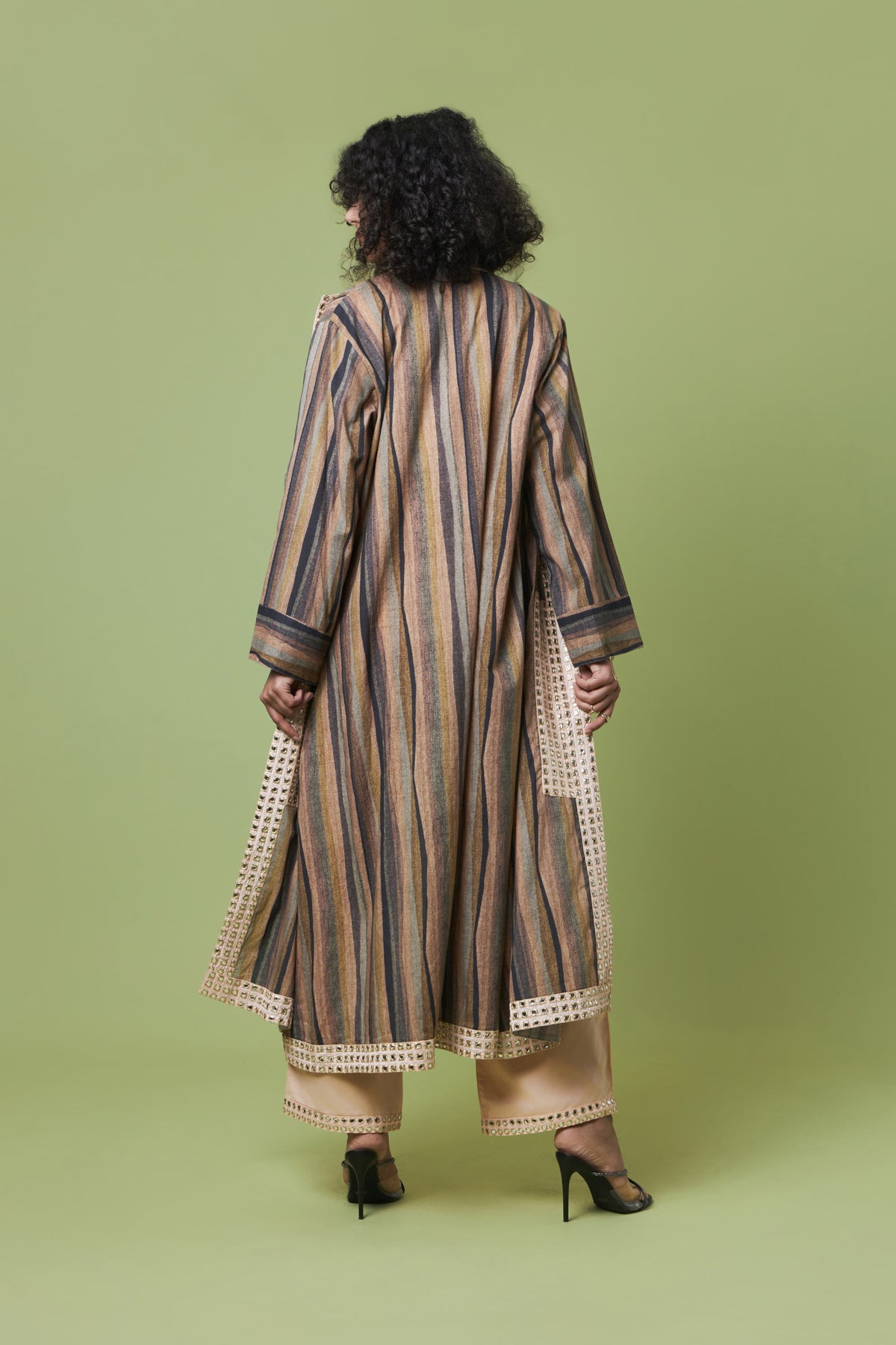 A woman viewed from behind wearing a long beige and brown vertically striped tunic with cream lace trim, paired with beige pants and black heels, against a solid light green studio background.