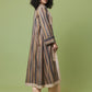 A woman modeling a long, loose-fitting striped tunic or kurta in beige, brown, and muted blue tones, paired with beige pants and black heels, set against a solid sage green studio background.