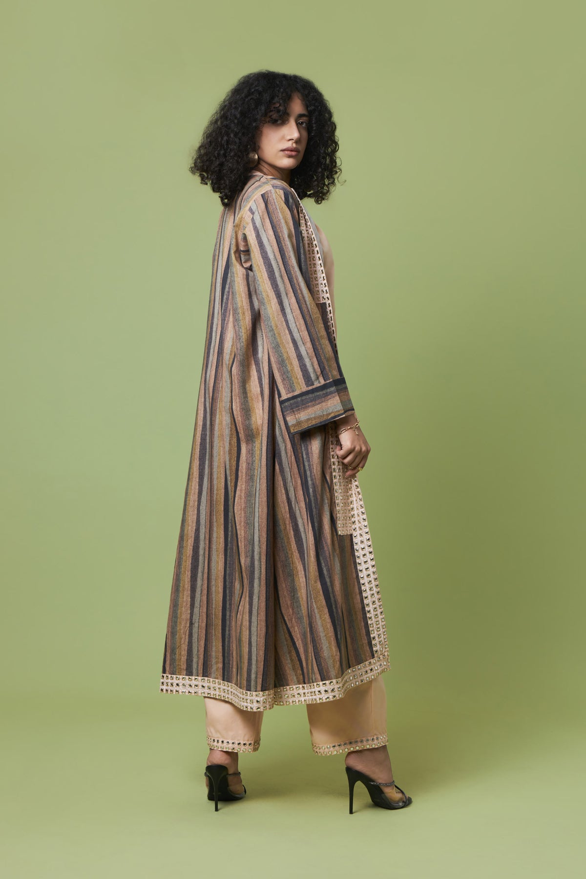 A woman modeling a long, loose-fitting striped tunic or kurta in beige, brown, and muted blue tones, paired with beige pants and black heels, set against a solid sage green studio background.