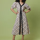 A woman modeling a knee-length midi dress with a beige and black trellis pattern, featuring a black V-neckline, black tie belt, and puff sleeves, set against a solid sage green background.