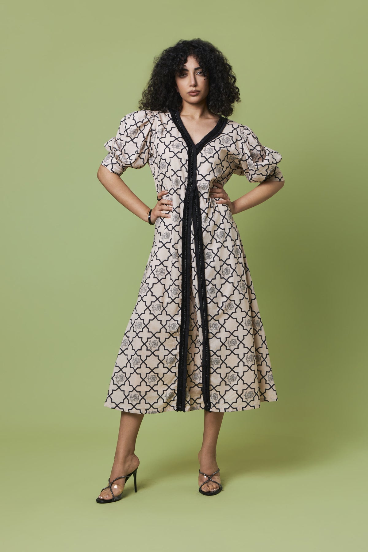A woman modeling a knee-length midi dress with a beige and black trellis pattern, featuring a black V-neckline, black tie belt, and puff sleeves, set against a solid sage green background.