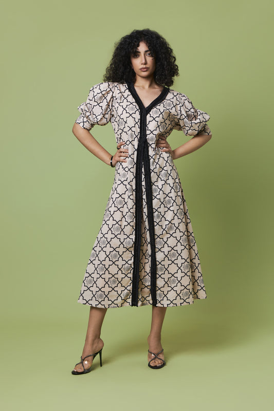 A woman modeling a knee-length midi dress with a beige and black trellis pattern, featuring a black V-neckline, black tie belt, and puff sleeves, set against a solid sage green background.