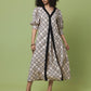 A woman modeling a midi-length dress with a beige and black geometric trellis pattern, featuring a black V-neckline and front trim, set against a solid light green studio background.