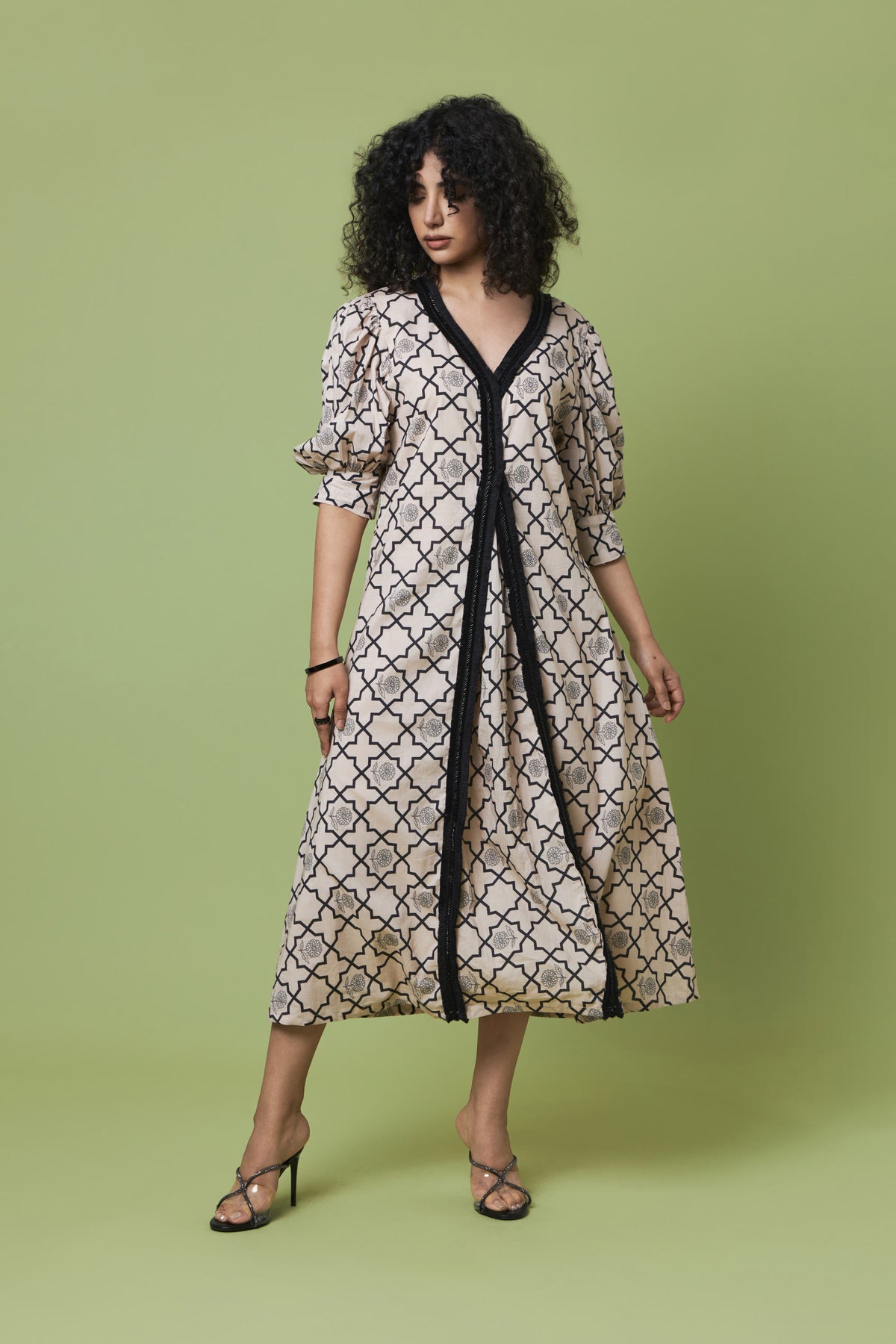 A woman modeling a midi-length dress with a beige and black geometric trellis pattern, featuring a black V-neckline and front trim, set against a solid light green studio background.