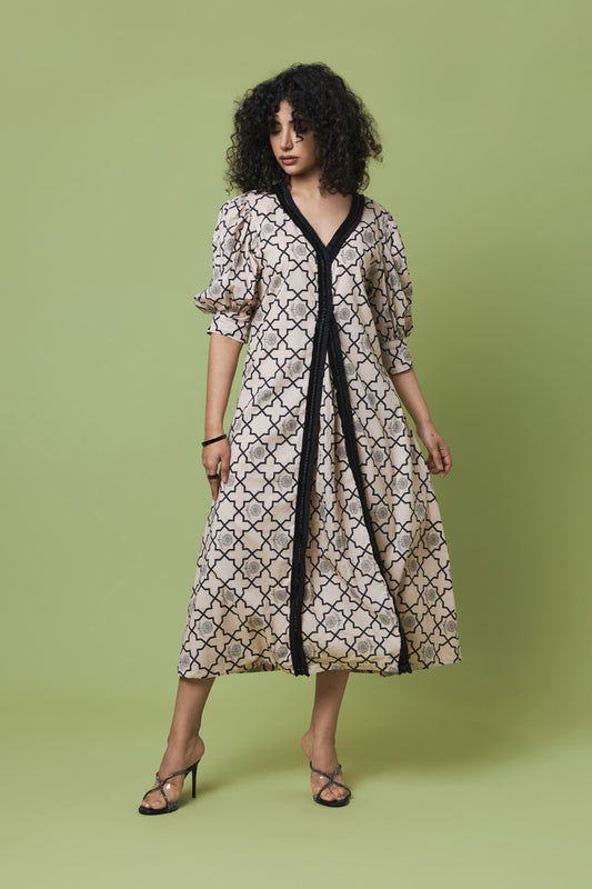 A woman modeling a midi-length dress with a beige and black geometric trellis pattern, featuring a black V-neckline and front trim, set against a solid light green studio background.