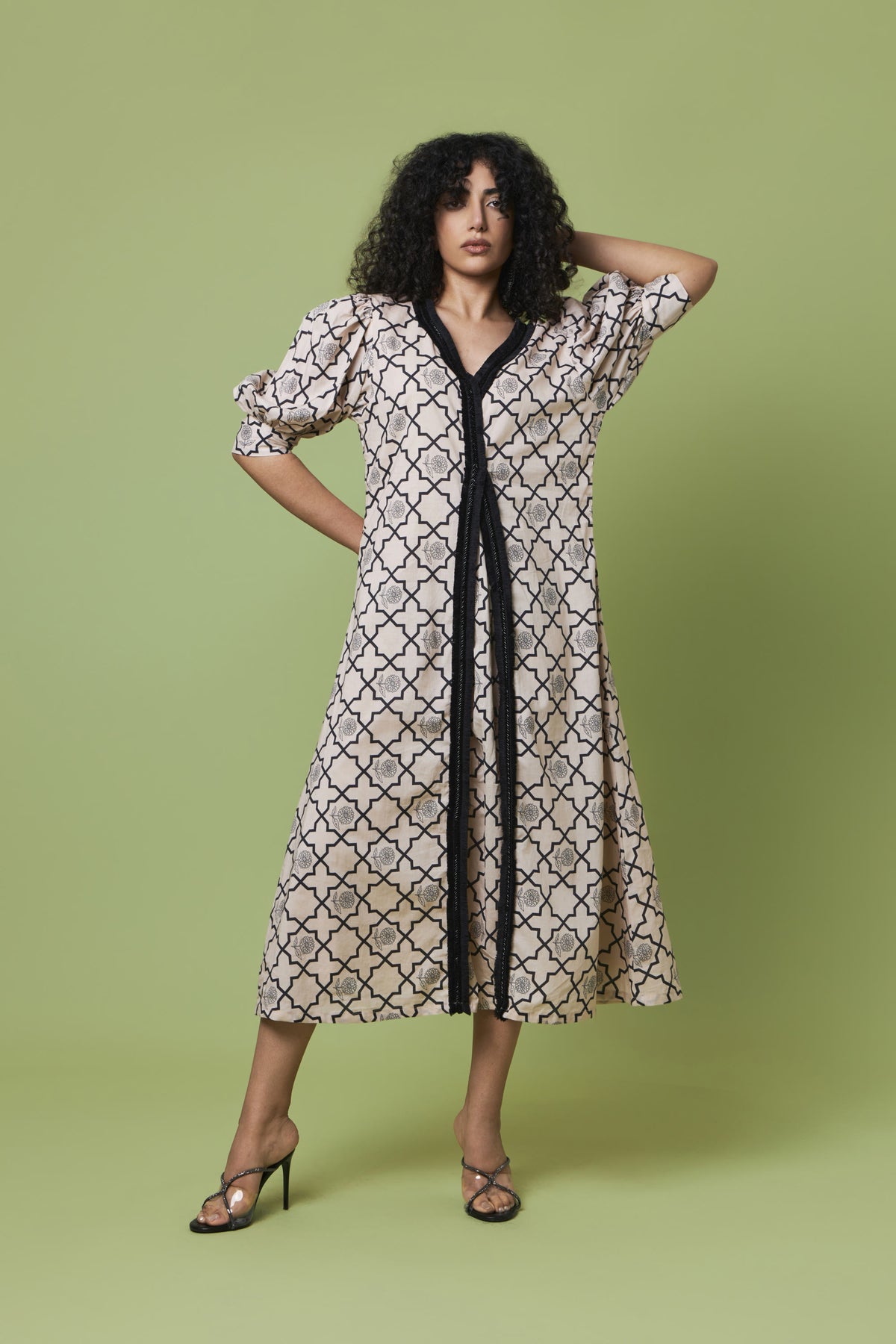 A woman modeling a knee-length patterned dress with puff sleeves and a black tie-front trim, standing against a solid light green studio background.
