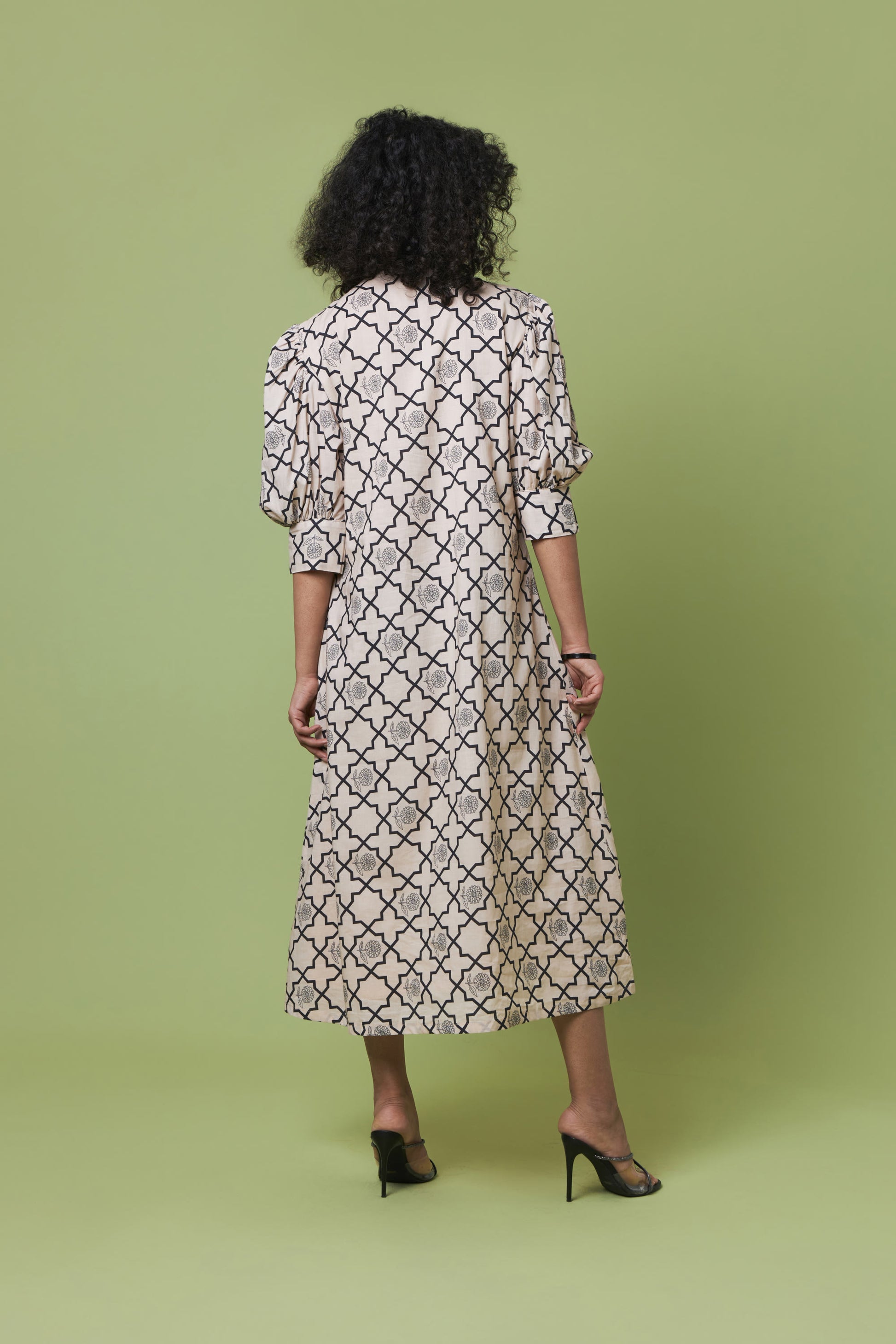 A woman viewed from behind wearing a long-sleeved midi dress with a black trellis pattern on a cream background, paired with black high heels, set against a solid light green studio backdrop.