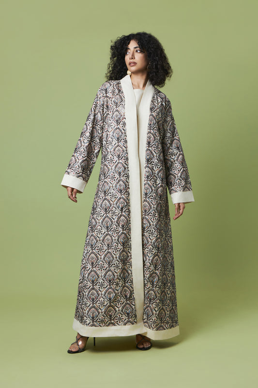 A woman modeling a long, open-front robe with a detailed black and gray damask pattern and a contrasting cream/off-white collar, cuffs, and hem, set against a solid light green studio background.