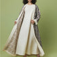 A woman stands against a solid sage green studio background wearing a long cream-colored dress with a matching open-front abaya and a patterned outer layer draped over her shoulders.