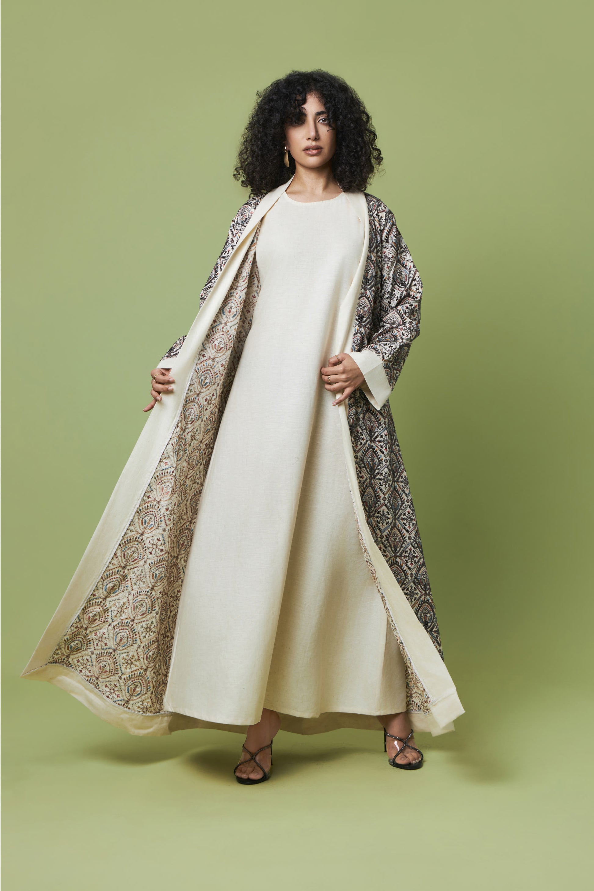 A woman stands against a solid sage green studio background wearing a long cream-colored dress with a matching open-front abaya and a patterned outer layer draped over her shoulders.