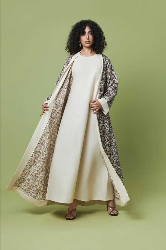 A woman stands against a solid sage green studio background wearing a long cream-colored dress with a matching open-front abaya and a patterned outer layer draped over her shoulders.