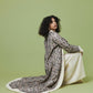 A woman with curly black hair sits on a light green studio floor wearing a long-sleeved cream and black paisley patterned shawl draped over a cream dress.