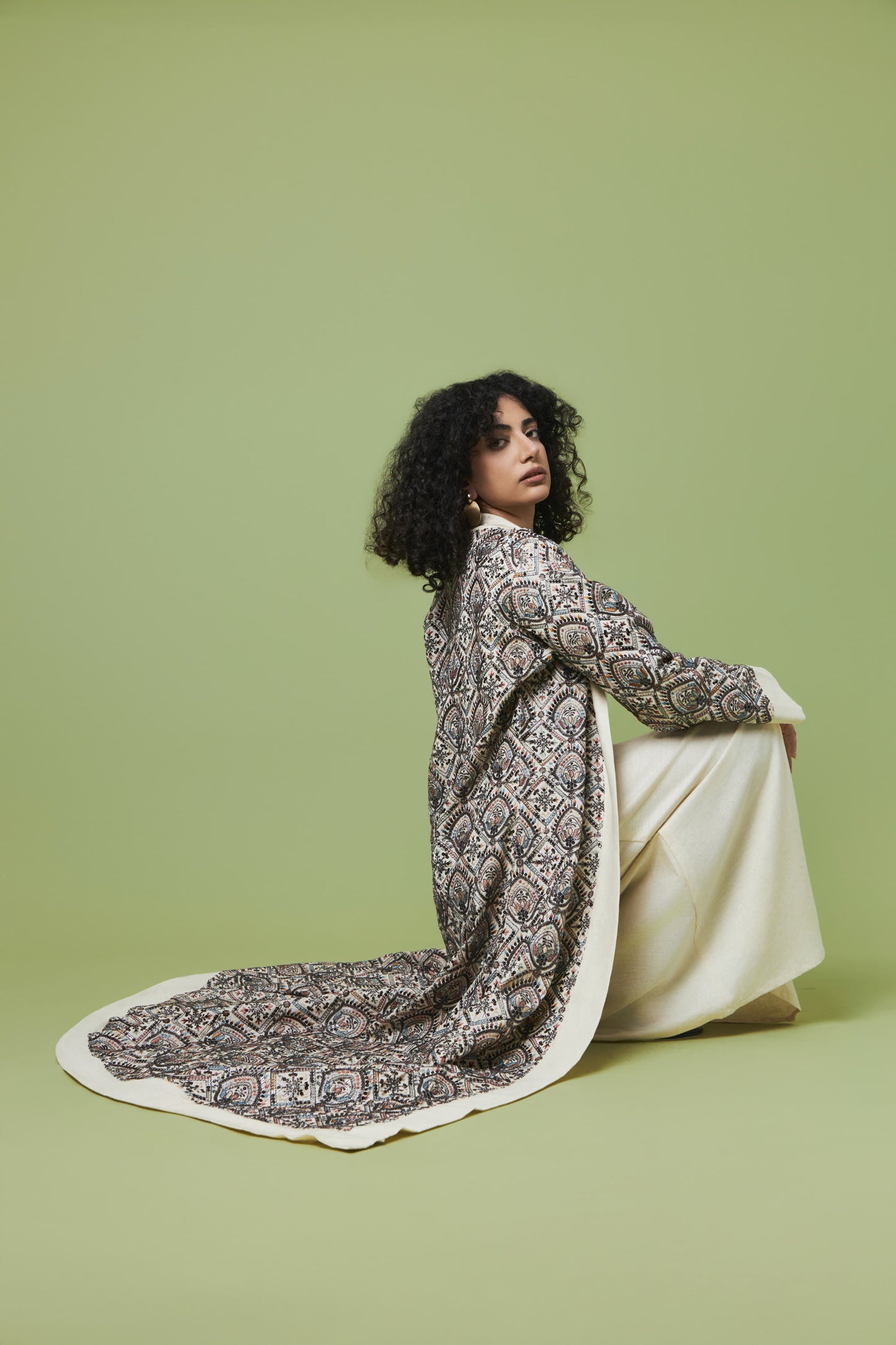 A woman with curly black hair sits on a light green studio floor wearing a long-sleeved cream and black paisley patterned shawl draped over a cream dress.