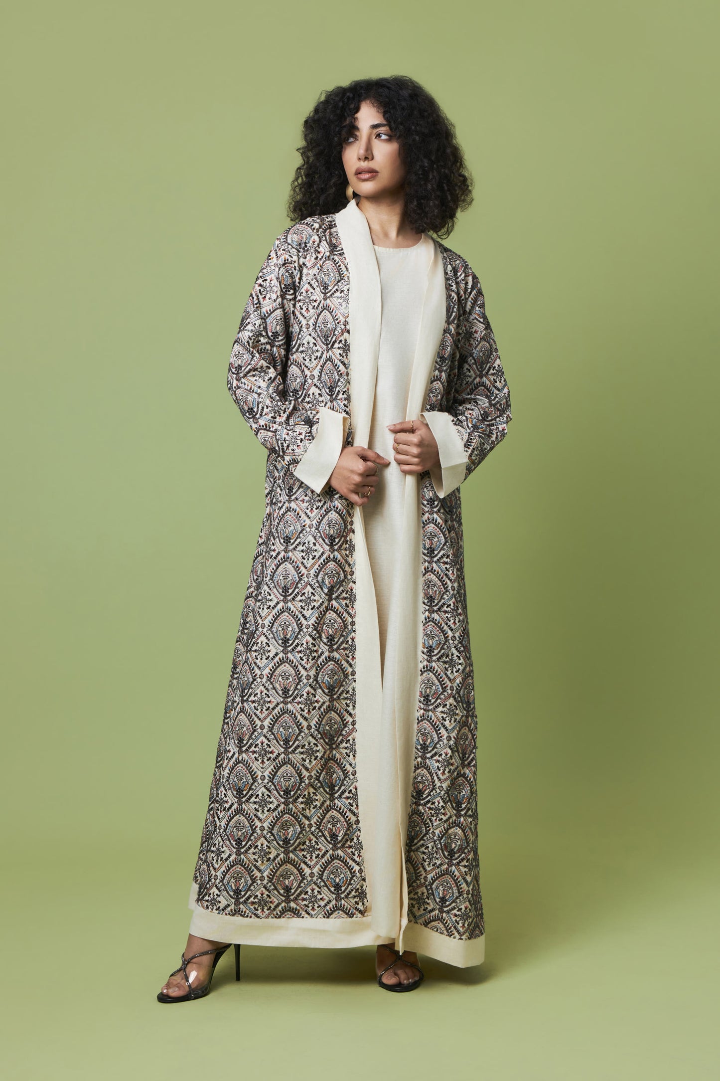 A woman modeling a long, open-front abaya with a detailed black and gray geometric pattern over a cream inner dress, set against a solid sage green studio background.