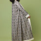 A woman modeling a long, flowing kaftan-style dress with an intricate black, gray, and beige floral pattern and solid cream borders, set against a solid sage green studio background.