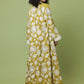 A woman stands with her back to the camera wearing a long-sleeved, floor-length yellow dress with a white leaf and floral pattern and a solid white hem, set against a solid light green studio background.