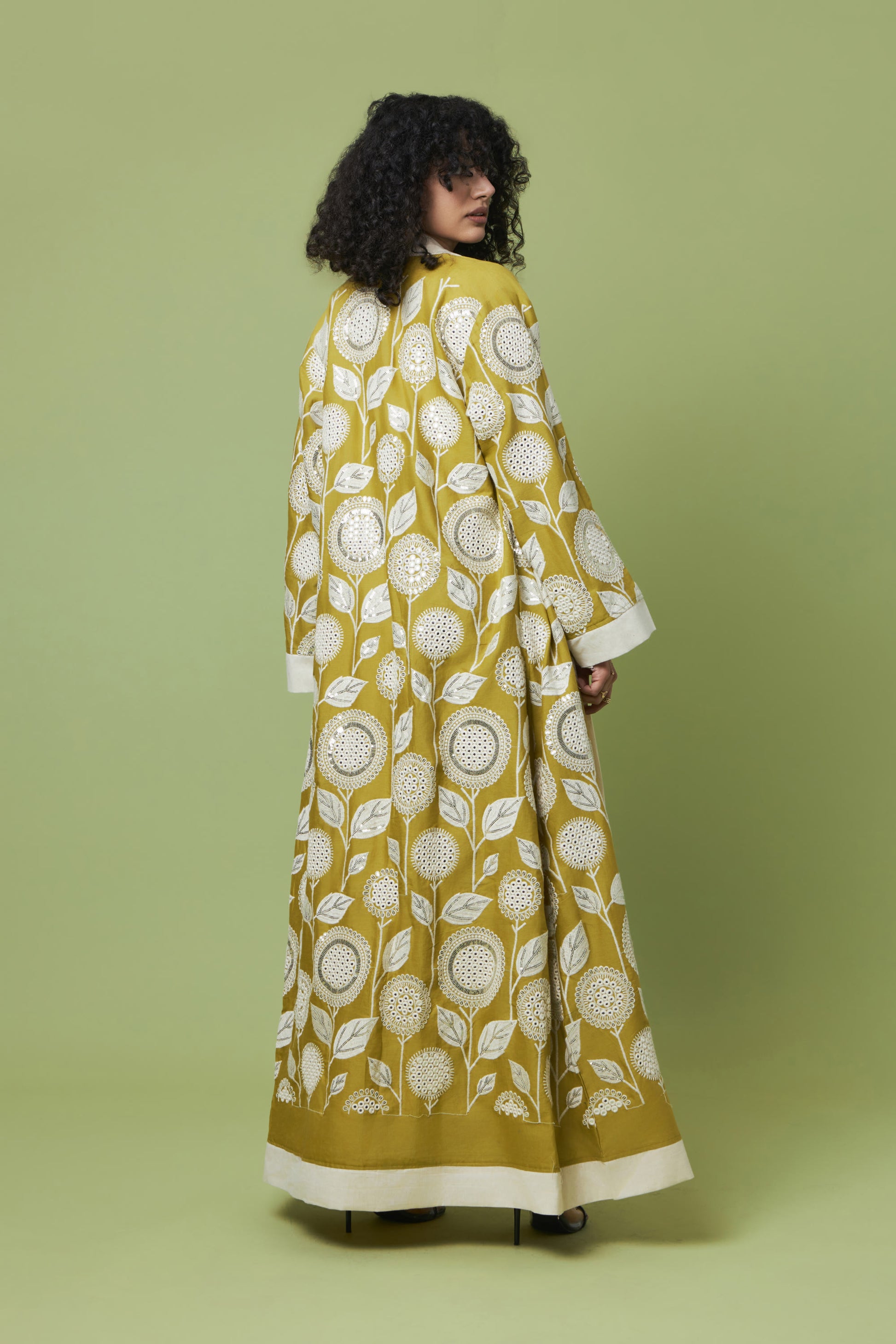 A woman stands with her back to the camera wearing a long-sleeved, floor-length yellow dress with a white leaf and floral pattern and a solid white hem, set against a solid light green studio background.