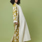A model with curly black hair is shown from the back wearing a long white coat over a mustard yellow and white patterned dress, set against a solid sage green studio background.