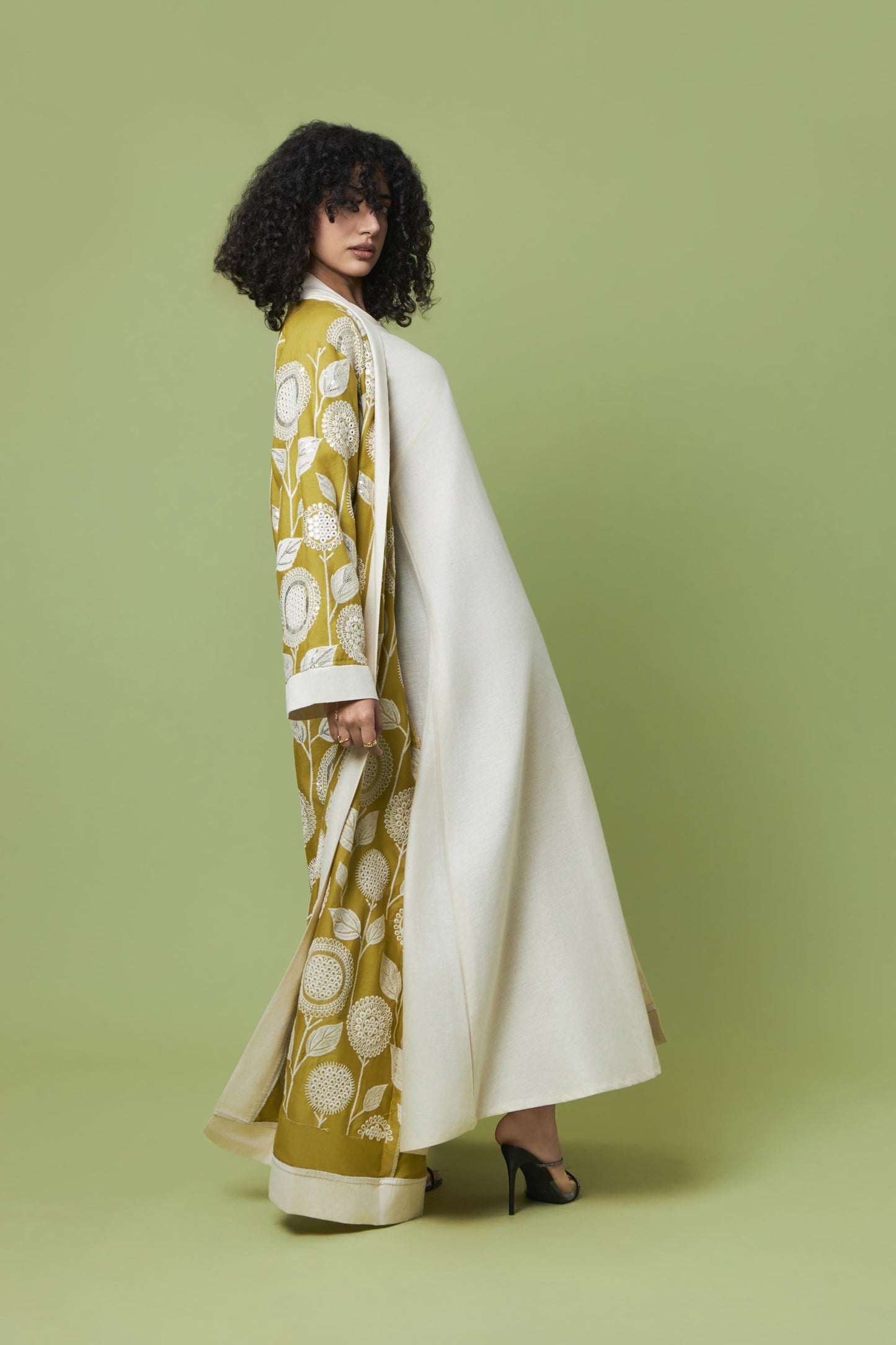 A model with curly black hair is shown from the back wearing a long white coat over a mustard yellow and white patterned dress, set against a solid sage green studio background.