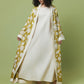 A woman stands against a solid sage green studio background wearing a long cream dress underneath an open, long-sleeved mustard yellow kimono-style coat with a white botanical pattern.