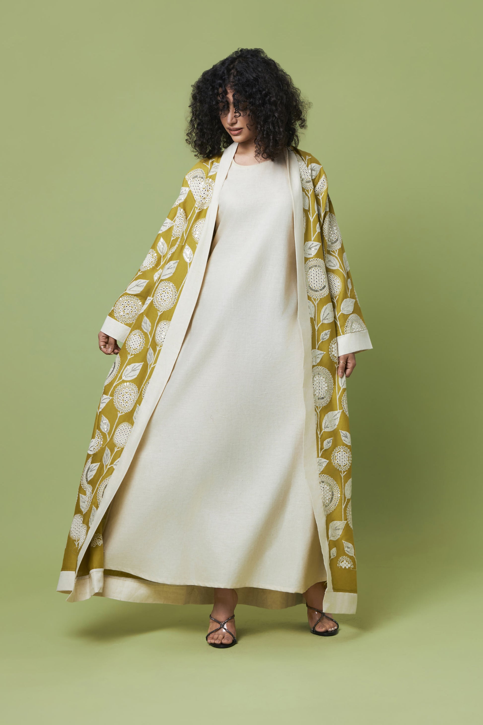 A woman stands against a solid sage green studio background wearing a long cream dress underneath an open, long-sleeved mustard yellow kimono-style coat with a white botanical pattern.