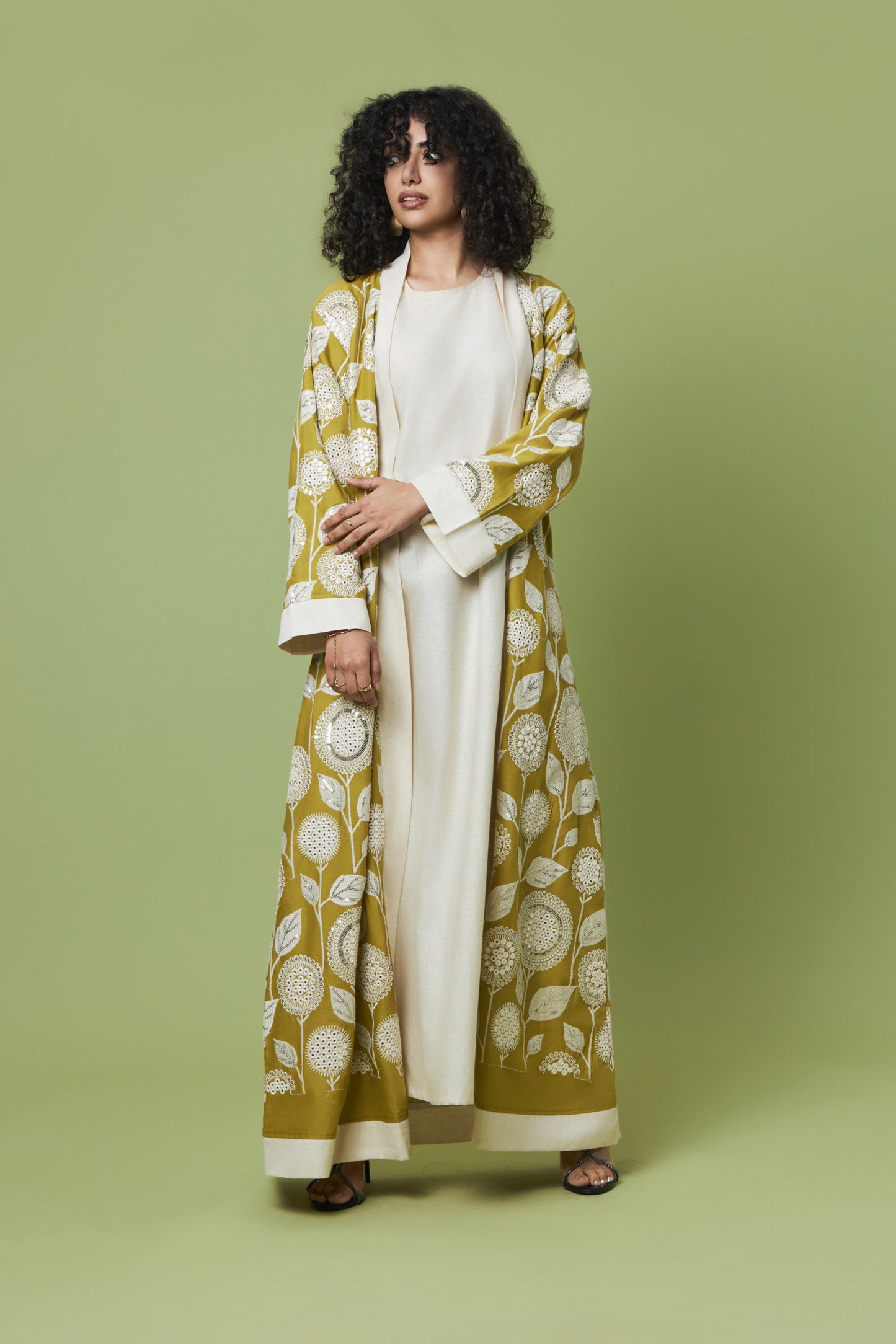 A woman modeling a long olive green and cream printed kaftan robe over a cream dress, standing against a solid olive green studio background.