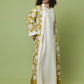 A woman modeling a long cream-colored dress with a matching open-front abaya featuring a mustard yellow and cream botanical pattern, set against a solid sage green studio background.