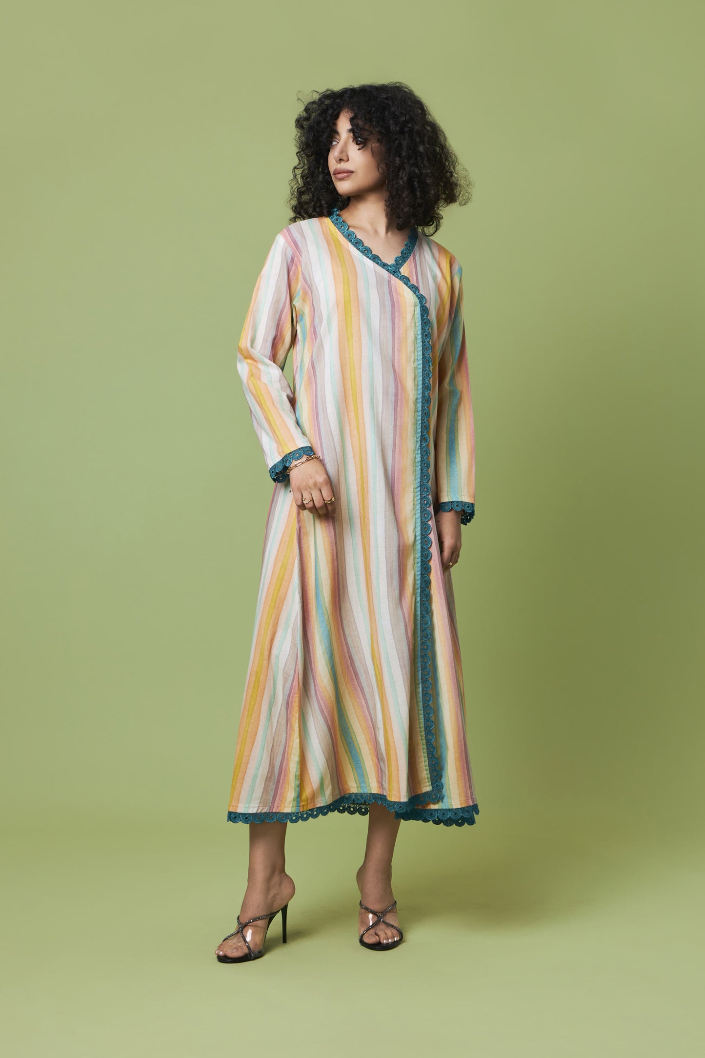 A woman modeling a long-sleeved, wrap-style maxi dress with vertical pastel stripes in pink, yellow, orange, and blue, featuring teal trim, set against a solid sage green studio background.